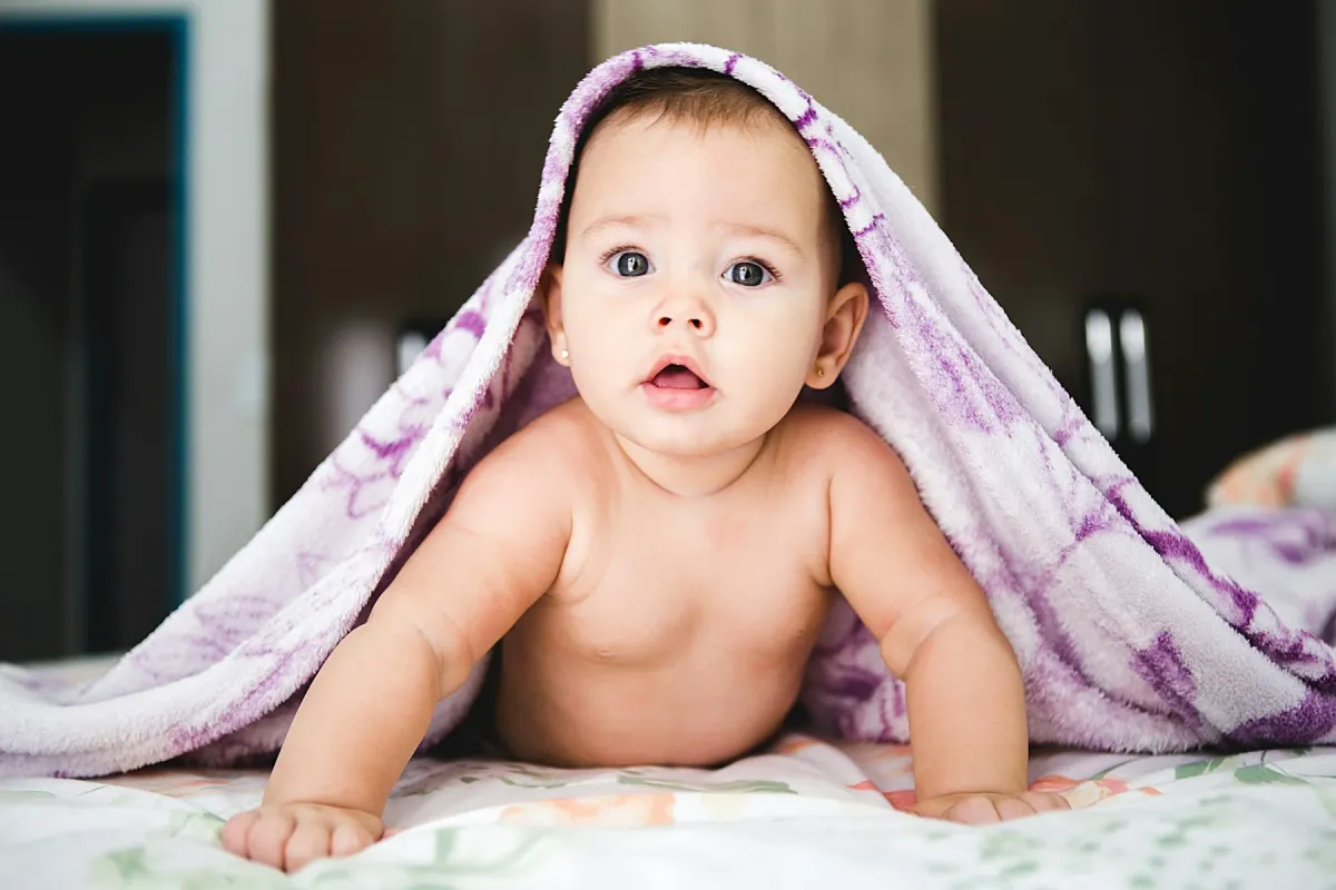 Bebê de barriga para baixo sobre a cama, olhando para a câmera sob uma manta lilás