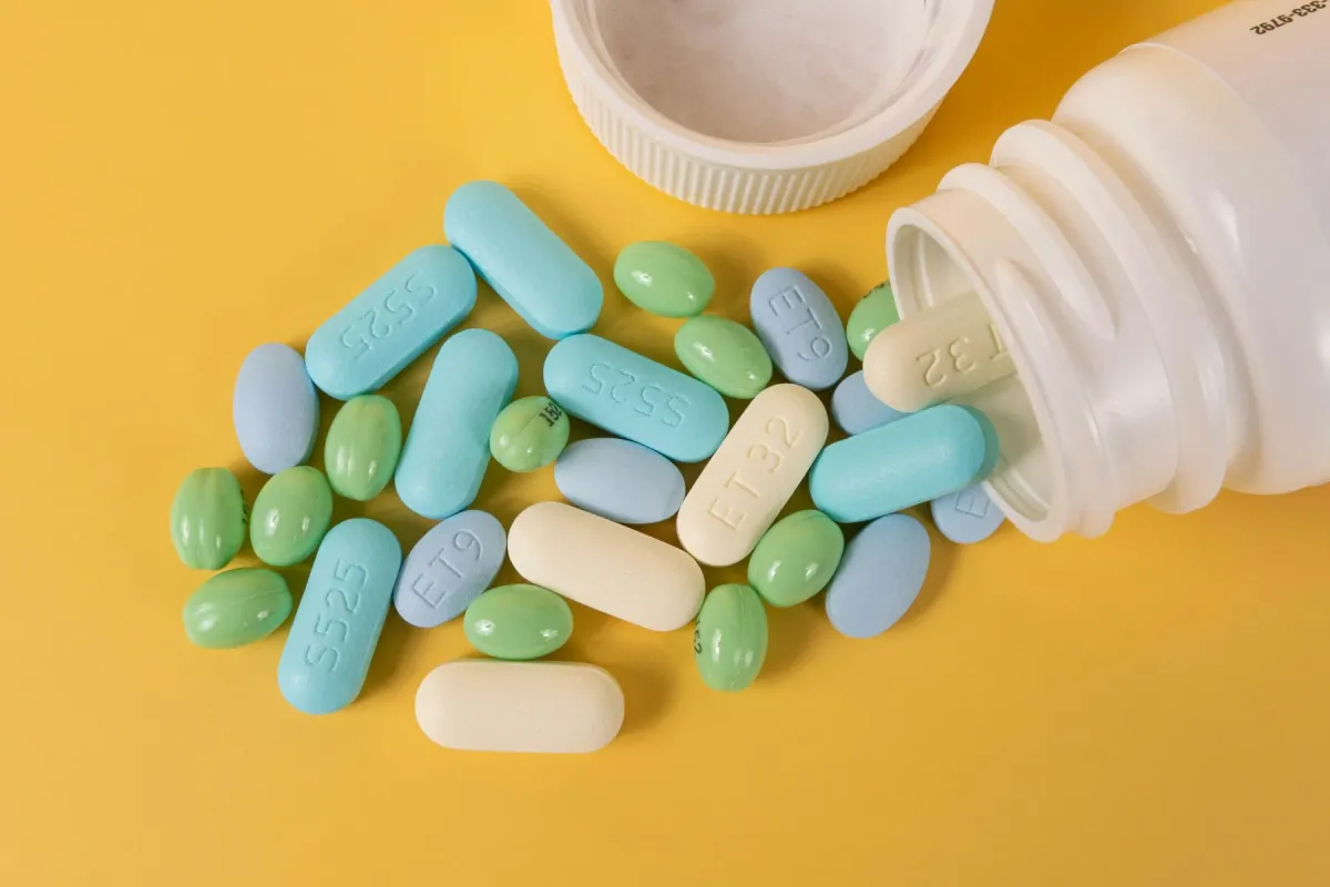 Comprimidos coloridos espalhados ao lado de um frasco de medicamento sobre fundo amarelo