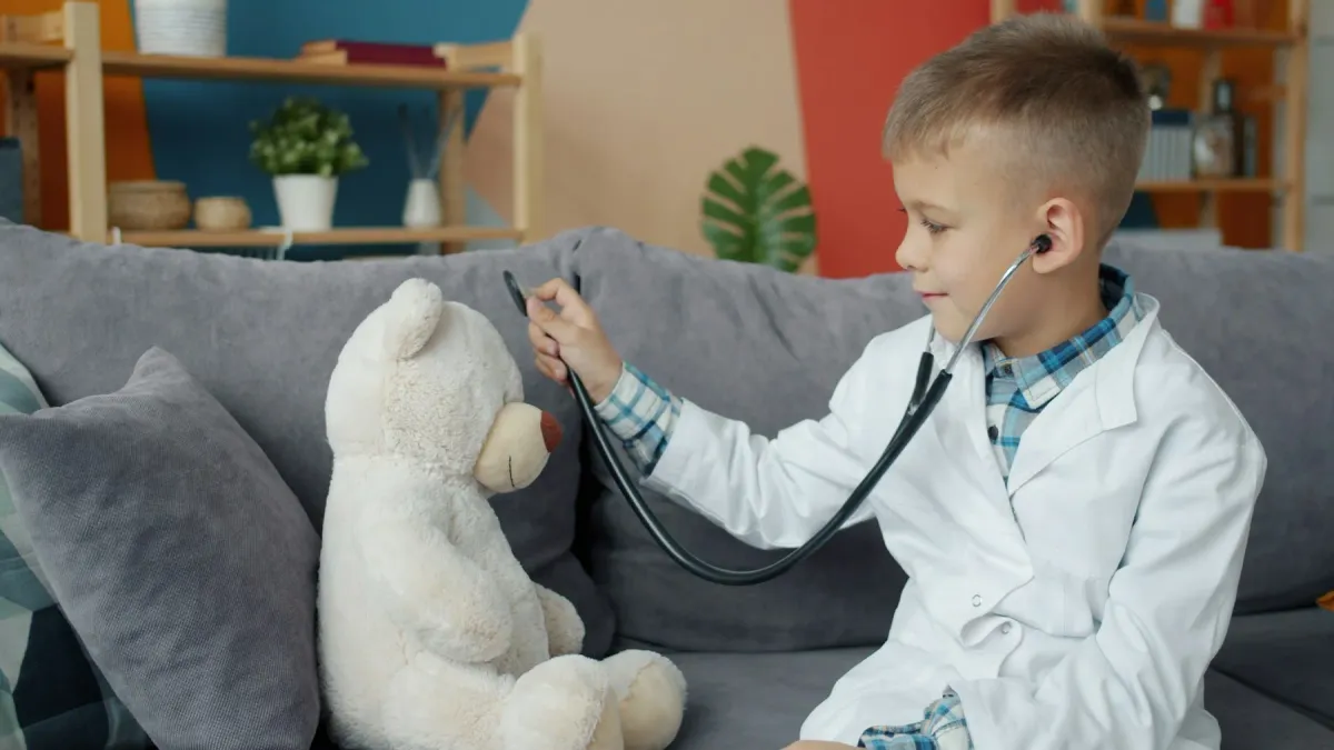 Menino de jaleco ausculta um urso de pelúcia com estetoscópio no sofá
