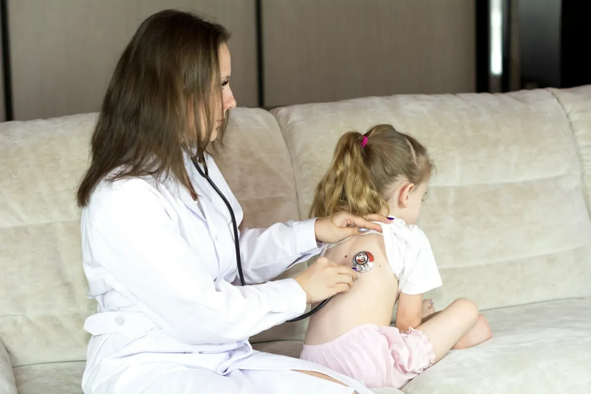 Médica de jaleco ausculta as costas de uma menina sentada no sofá