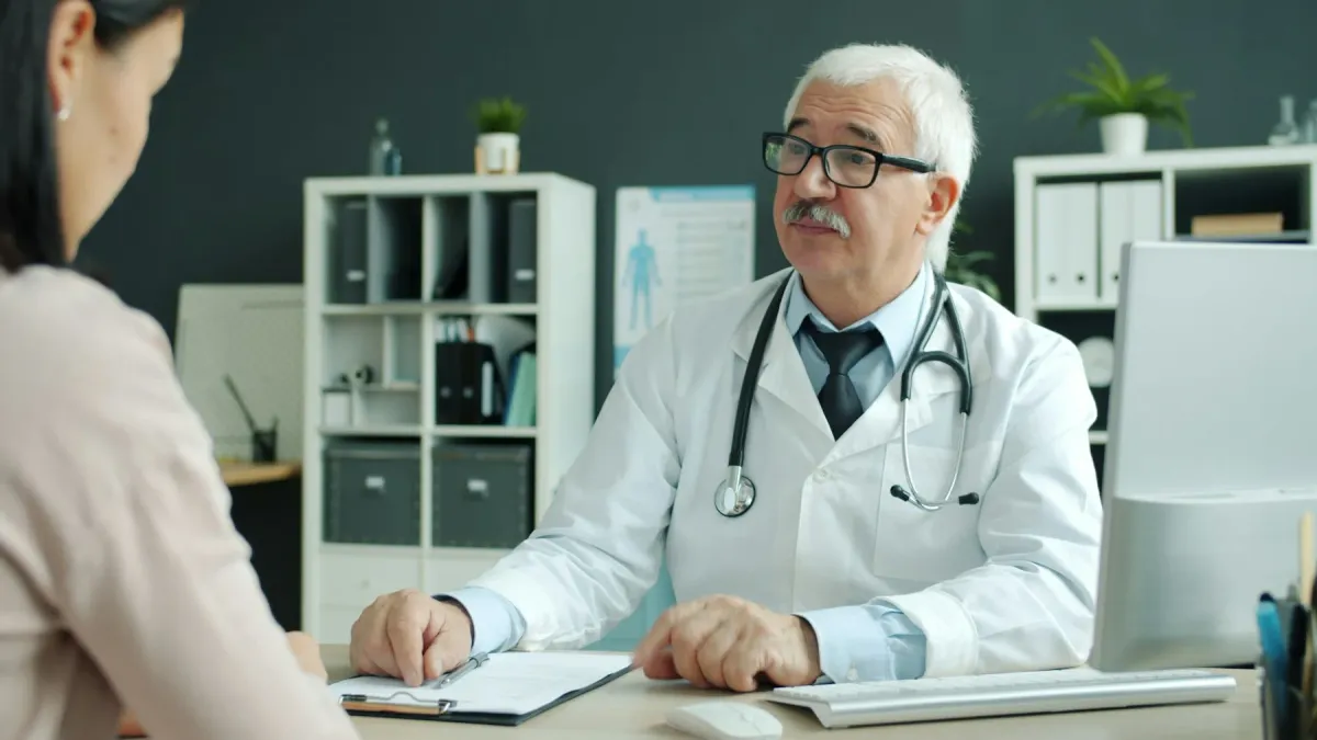 Médico idoso de jaleco conversa com uma paciente em consultório