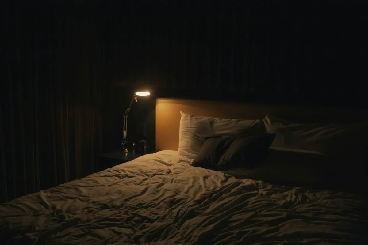 Cama de casal em quarto escuro com um abajur aceso na mesa de cabeceira