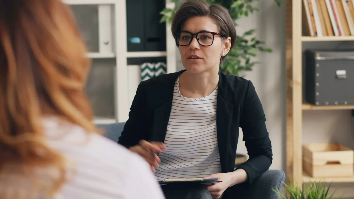Psicóloga de óculos com prancheta conversa com uma paciente em consultório