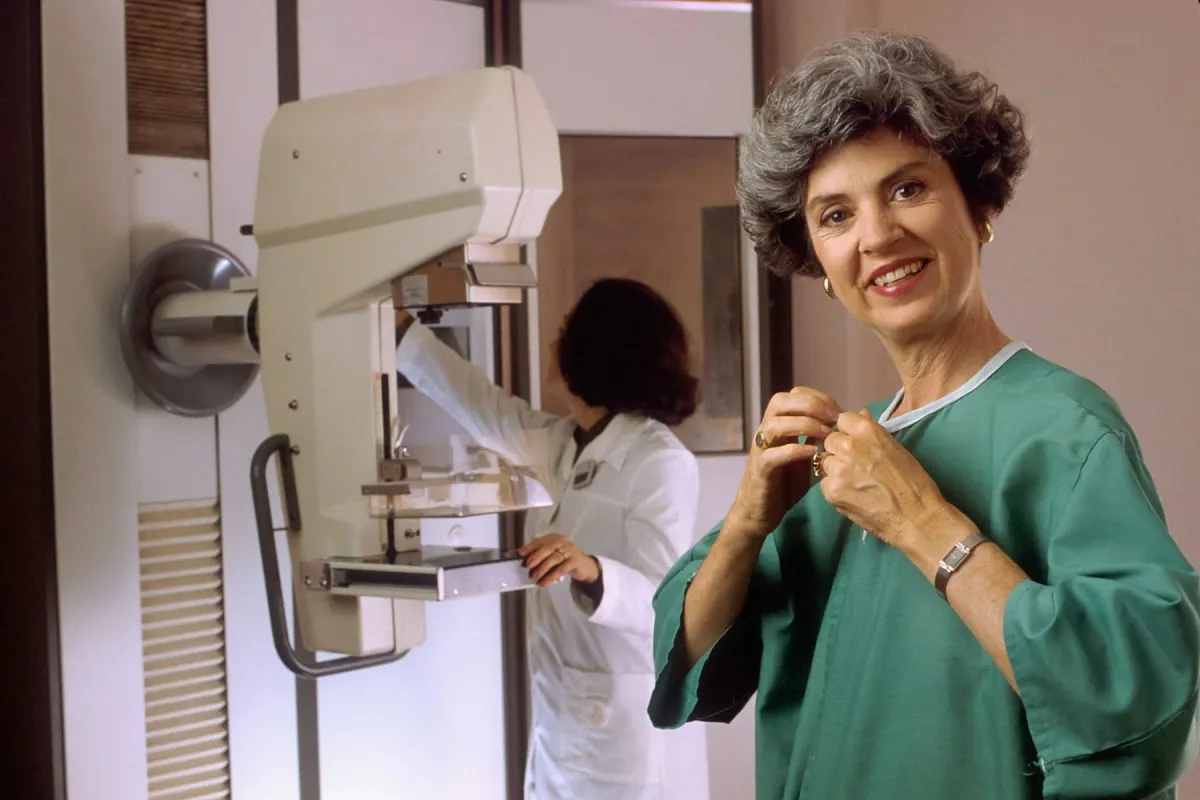 Mulher sorri ao lado de um mamógrafo enquanto uma profissional ajusta o equipamento