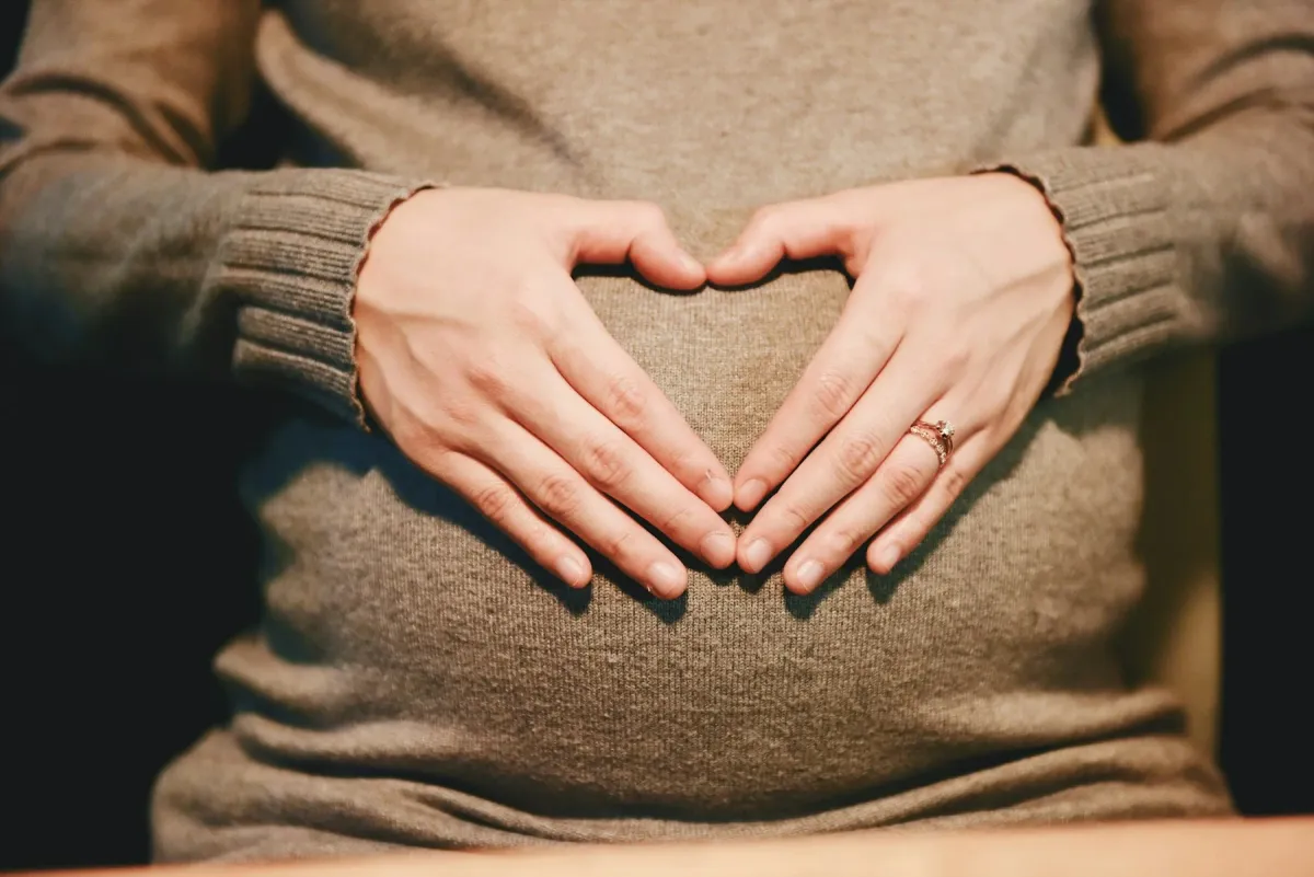 Gestante de suéter faz um coração com as mãos sobre a barriga