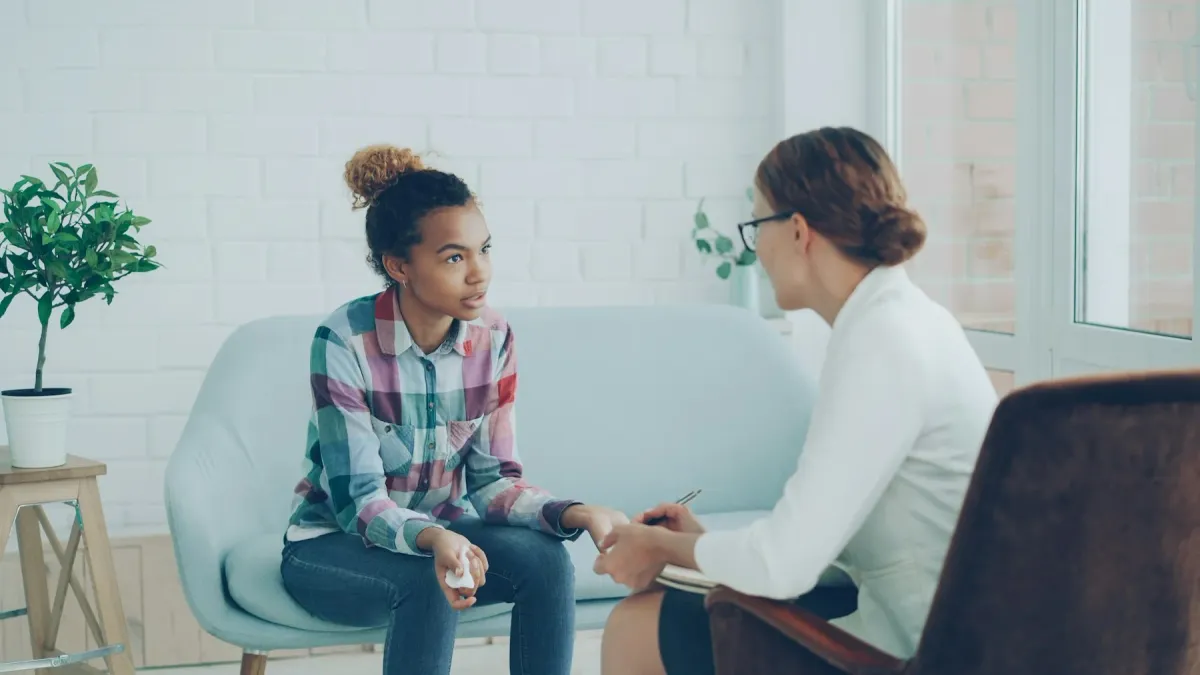 Terapeuta com prancheta conversa com uma jovem sentada em um sofá azul