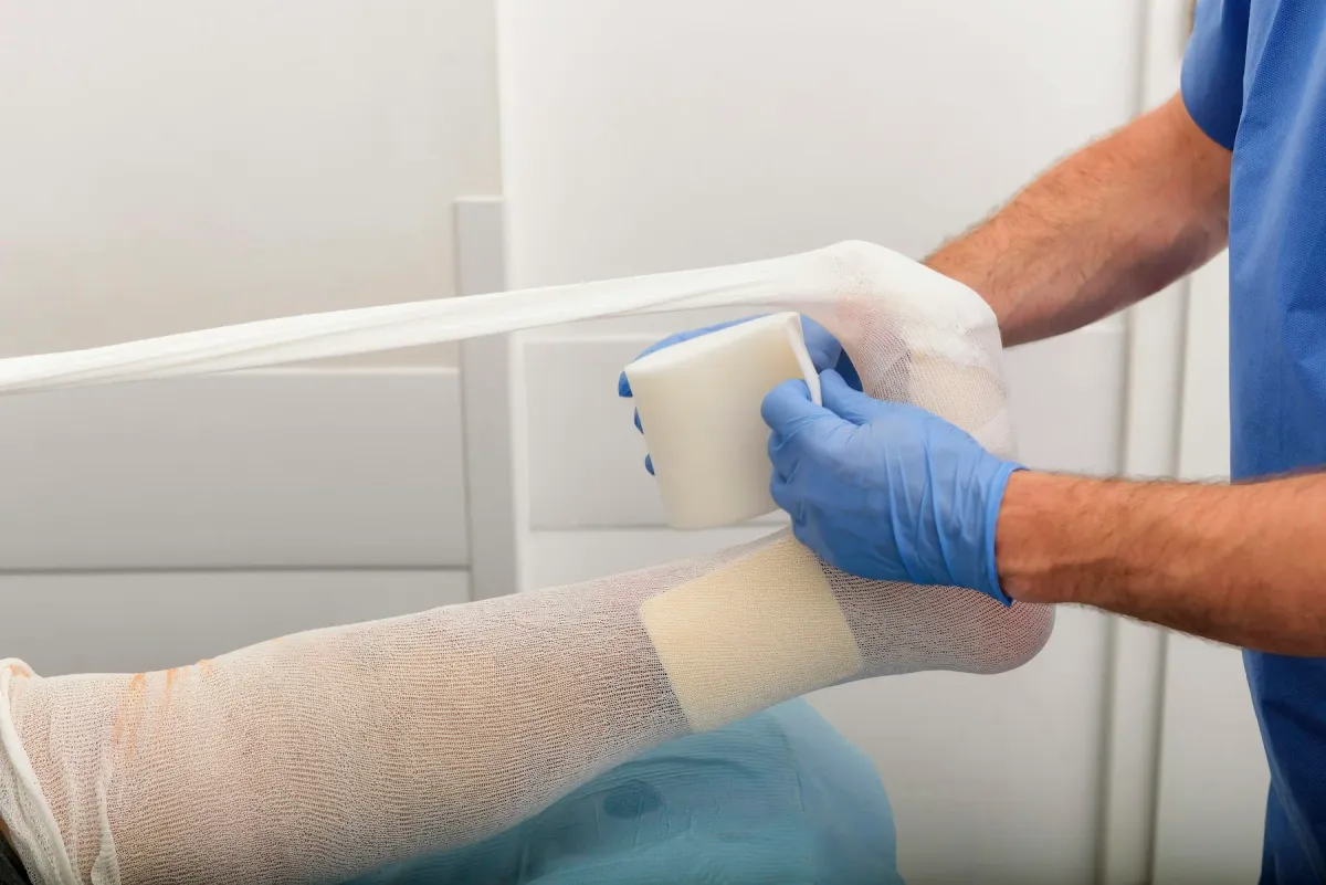 Profissional de luvas azuis aplica curativo com atadura na perna de um paciente