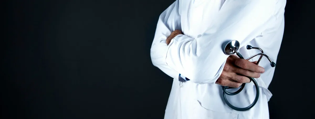 Médico de jaleco branco e braços cruzados segura um estetoscópio, em fundo escuro