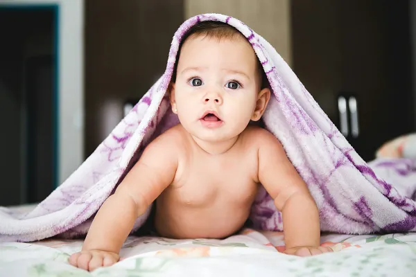 Bebê de barriga para baixo sobre a cama, olhando para a câmera sob uma manta lilás