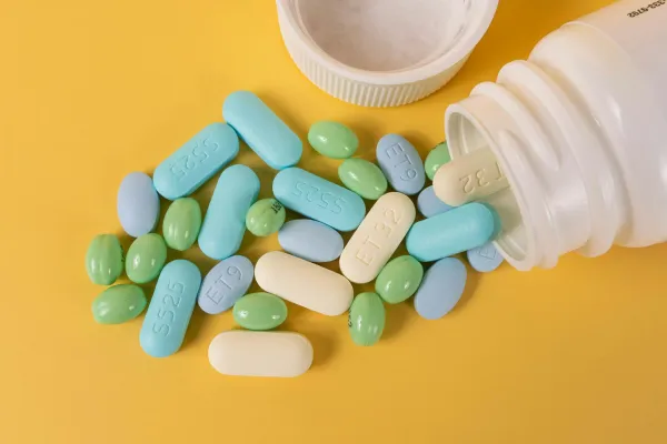 Comprimidos coloridos espalhados ao lado de um frasco de medicamento sobre fundo amarelo