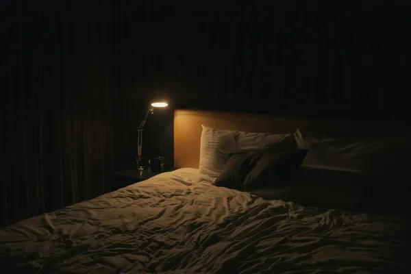 Cama de casal em quarto escuro com um abajur aceso na mesa de cabeceira