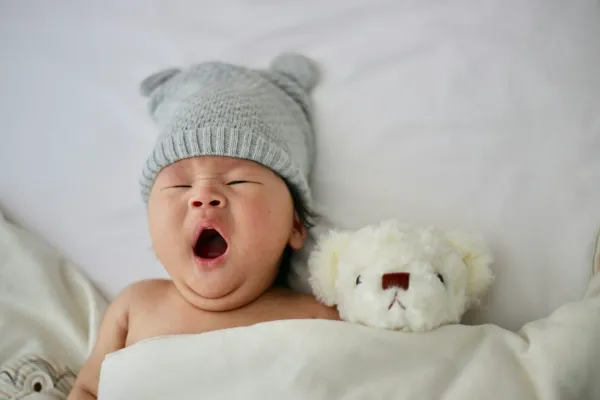 Recém-nascido de gorro cinza bocejando ao lado de um ursinho de pelúcia