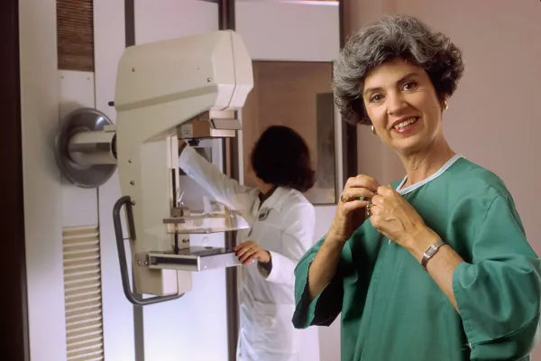 Mulher sorri ao lado de um mamógrafo enquanto uma profissional ajusta o equipamento