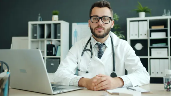 Médico de jaleco e estetoscópio sentado à mesa do consultório com notebook e anotações