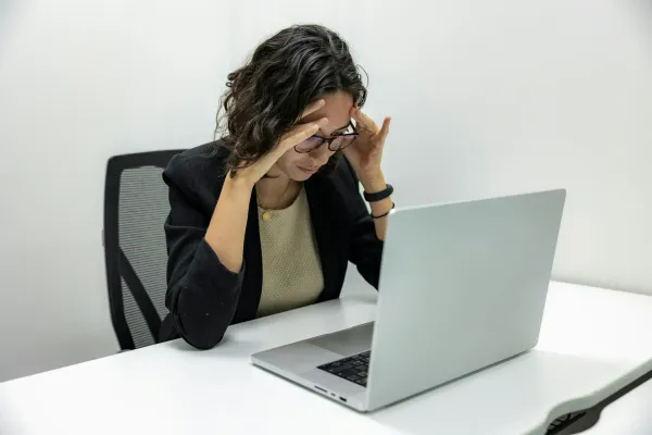 Mulher com as mãos nas têmporas em frente ao notebook, com dor de cabeça