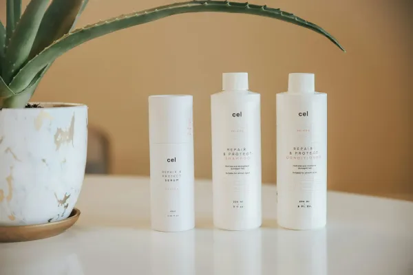 Três frascos brancos de shampoo, condicionador e sérum sobre uma mesa ao lado de uma planta
