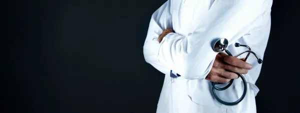 Médico de jaleco branco e braços cruzados segura um estetoscópio, em fundo escuro